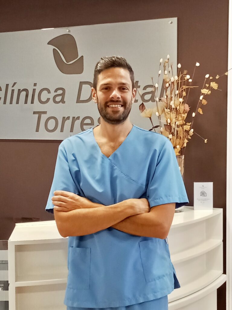 Hombre sonriente en uniforme azul de odontología, de pie con los brazos cruzados frente a la Clínica Dental Torres, con decoración moderna en el fondo.