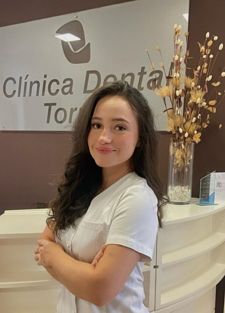 Mujer sonriente con cabello largo y oscuro, vestida con uniforme blanco, de pie frente al mostrador de la Clínica Dental Torremolinos, con un letrero de la clínica en el fondo y decoración floral a su lado.